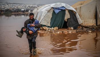 سوري يحمل طفلاً أثناء إخلاء متعلقاته من الخيام التي غمرتها المياه في مخيم كفر لوسين للاجئين في إدلب